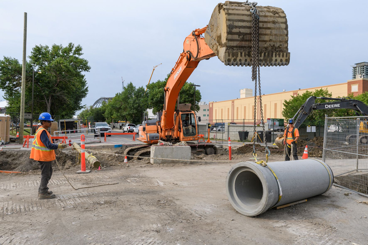 An excavator gets ready to lift a pipe with assistance from two construction workers in Beltline East.