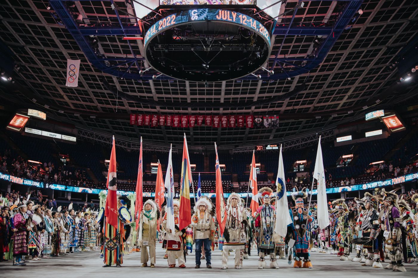 inside an arena on the arena floor, Aboriginal folks are dressed in traditional wear.  there is a line of 7 people holding flags centre of the image